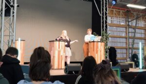 Brie und Amelie stehen auf der Bühne. Brie beschreibt ihren Weg in die Welt der BNE.